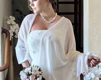 Puede incluir: Un chal de novia blanco con un delicado patrón de punto, usado sobre un vestido de novia blanco sin tirantes. La novia lleva un ramo de flores rosas y blancas.