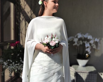 Puede incluir: Una novia con un vestido de novia blanco, una chalina blanca y un ramo de flores rosas y blancas. Está de pie frente a una ventana y una silla de mimbre blanca.