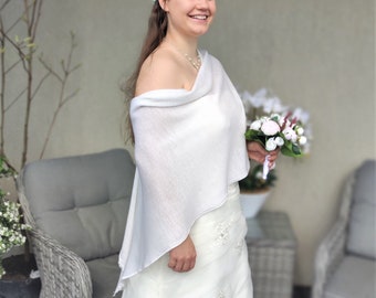 Puede incluir: Una novia con un vestido de novia blanco y un chal sonríe, sosteniendo un ramo de flores blancas. El vestido tiene una falda en capas con detalles bordados. Lleva un collar y un accesorio para el pelo.