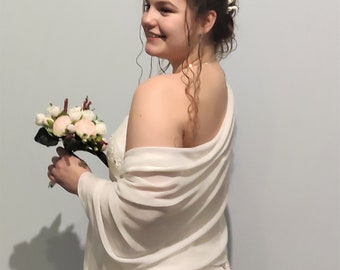 Puede incluir: Una estola de novia blanca drapeada sobre los hombros de una novia. La estola está hecha de una tela transparente y tiene un diseño sencillo. La novia lleva un vestido de novia blanco y tiene una corona de flores en el pelo.