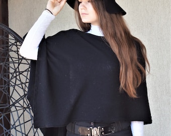 May include: A black poncho with a white trim around the neckline. The poncho is worn over a white turtleneck and black trousers with a black belt. The person is wearing a black hat with a white band.