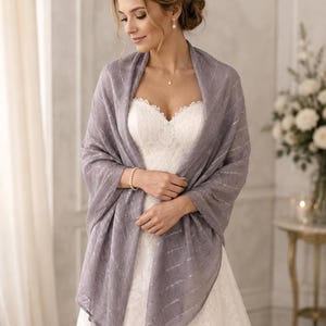 Puede incluir: Un chal de color lavanda con un sutil patrón de rayas plateadas metálicas, drapeado sobre un vestido blanco sin tirantes. La tela ligera tiene una textura suave y forma rectangular, proporcionando cobertura sobre los hombros y los brazos.