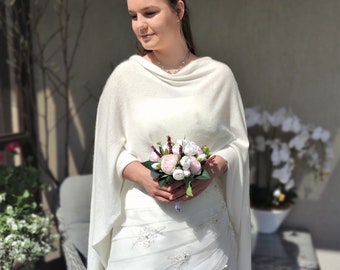 Puede incluir: Una novia con un vestido de novia blanco y un chal sostiene un ramo de flores rosas y blancas. El vestido tiene adornos florales. El chal está drapeado sobre los hombros. El fondo incluye un patio y plantas.