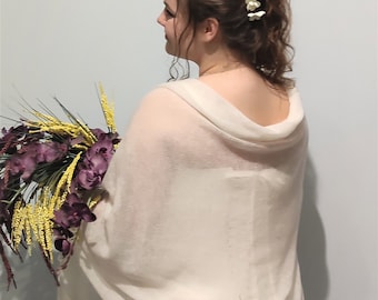 May include: A woman wearing a cream-colored shawl and a white dress, holding a bouquet of purple and yellow flowers. White flower hair accessories adorn her hair. The shawl drapes over her shoulders.