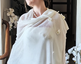 May include: A cream-colored wedding shawl with delicate floral embellishments. The shawl is draped over a white dress, and the image is taken in natural light. The overall aesthetic is soft and romantic, suitable for bridal wear.