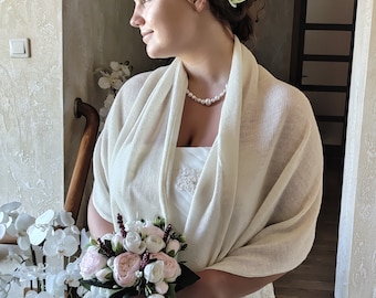 May include: A bride wearing a white wedding dress and a cream-colored shawl. She holds a bouquet of pink and white flowers. A pearl necklace and a flower in her hair complete the look. The setting appears to be indoors.