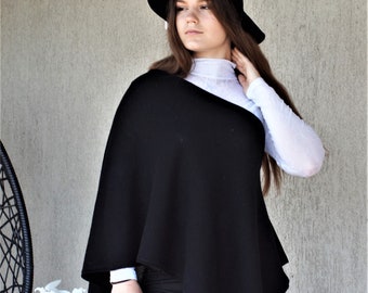 May include: A black knit poncho with a long, asymmetrical hemline. The poncho is worn over a white turtleneck shirt and black trousers. The person is wearing a black hat with a white band.