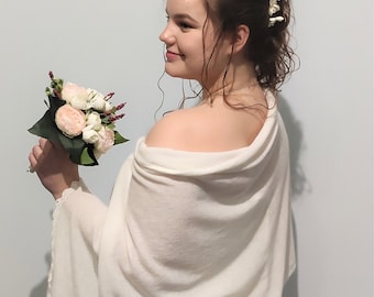Puede incluir: Un chal de novia blanco con un delicado borde de encaje. El chal está drapeado sobre los hombros de una persona que sostiene un ramo de flores blancas y rosas.