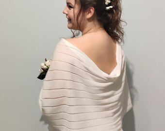 Puede incluir: Un chal de novia blanco con un delicado patrón de punto. El chal está drapeado sobre los hombros de una persona que lleva un vestido blanco y una diadema floral.