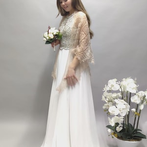 Pode incluir: Um vestido de noiva branco com um corpete de contas e um xale de renda transparente. O vestido tem uma saia fluida e é combinado com um buquê de flores brancas.