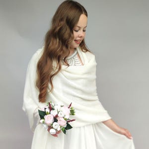 Peut inclure: Un châle tricoté blanc cassé doux drapé sur les épaules, associé à une robe blanche et un petit bouquet de fleurs artificielles roses et blanches avec des feuilles vertes. Le châle présente un motif en maille fine et texturée.