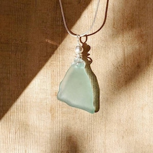 May include: A sea glass pendant necklace. The pendant is a pale blue, irregularly shaped piece of sea glass. It hangs from a silver chain with a small silver bead. The background is a wooden surface.
