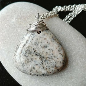 May include: A handcrafted pendant necklace featuring a smooth, speckled stone in shades of gray and cream. The stone is wire-wrapped with silver-colored metal and suspended from a delicate chain. The pendant is approximately 1.5 inches long.