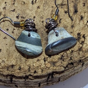 May include: Handmade earrings featuring natural stone pendants with gray and white stripes and dark gray accents. The stones are wire-wrapped with dark metal wire and accented with small yellow beads. The earrings have a simple hook design.