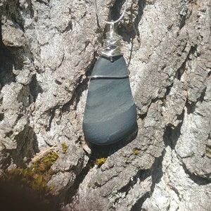 Peut inclure: Un collier pendentif en pierre gris foncé avec un fil d'argent et une chaîne. La pierre a une surface lisse et polie avec des bandes horizontales de gris plus clair. Le pendentif est présenté sur un fond d'écorce d'arbre texturée.