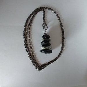 Puede incluir: Un collar con un colgante de piedras apiladas y pulidas en tonos negro, gris y blanco. El colgante cuelga de una cadena de metal oscuro. El collar se muestra sobre un fondo blanco.
