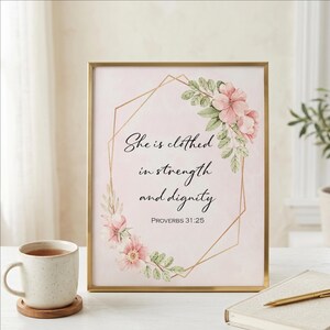 May include: A framed print with the quote "She is clothed in strength and dignity" in black script, accented by pink floral illustrations and a gold geometric frame. A mug of tea and a notebook are in the foreground.