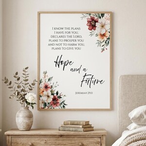 May include: A framed print with the text "I know the plans I have for you... Hope and a Future" and floral accents. The artwork features a light wood frame, with a white background and elegant script. The floral arrangement includes burgundy, peach, and white flowers.