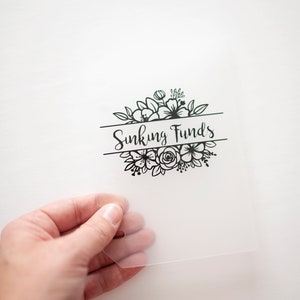 以下が含まれることがあります： 黒い花のデザインと「Sinking Funds」というテキストが印刷された透明なプラスチックシートです。
