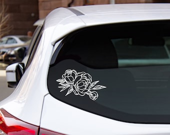 May include: White floral decal featuring a peony design on a car window. The decal shows a detailed outline of peony blooms and leaves, creating a delicate and elegant aesthetic. The car is white.