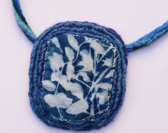 May include: A blue and white fabric pendant necklace with a floral design. The pendant is made of a woven fabric with a blue border and a white fabric center with a blue floral pattern.