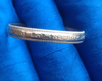 May include: A silver bangle bracelet with a decorative design. The bracelet is on a blue fabric background.