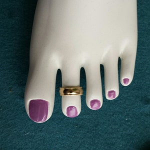 May include: A gold toe ring with a textured design. The ring is worn on the toe of a mannequin with purple nail polish.