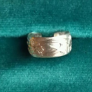May include: A silver toe ring with a floral design. The ring is on a teal green velvet surface.