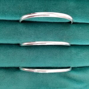 May include: Three silver bangle bracelets of varying sizes, stacked on a green velvet surface.