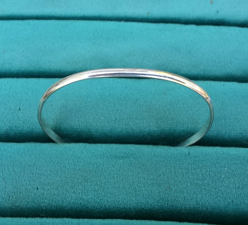 May include: A simple silver bangle bracelet, shown against a teal green background.