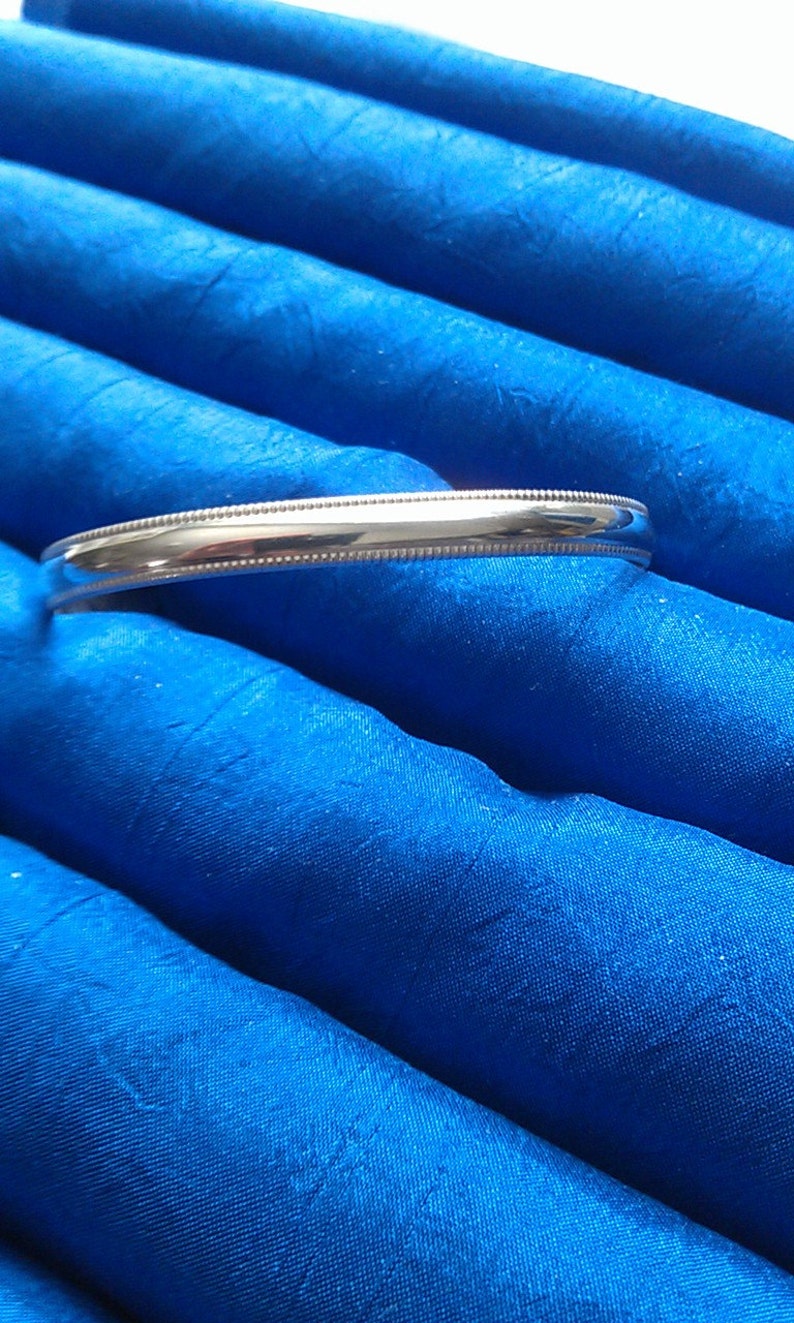 Pu&ograve; includere: Un braccialetto rigido in argento con un bordo perlato. Il braccialetto &egrave; su uno sfondo di tessuto blu.