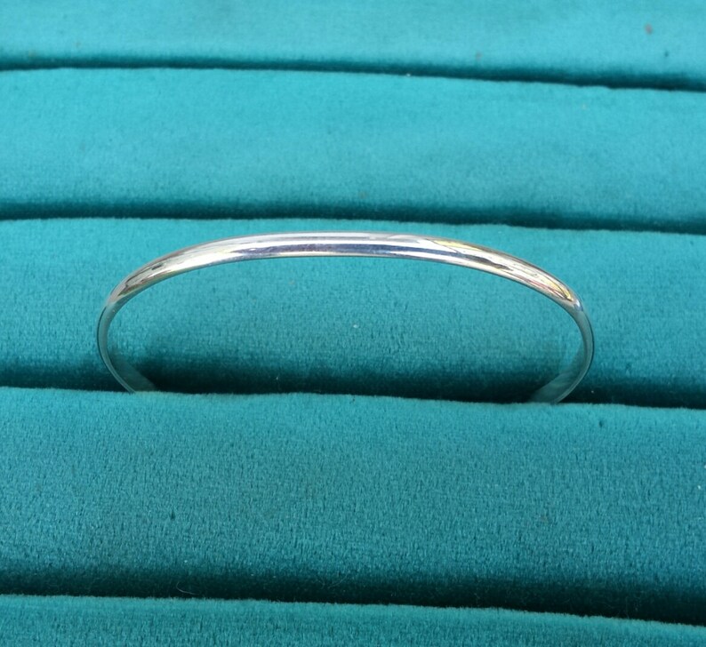 May include: A simple silver bangle bracelet, shown on a teal velvet background.