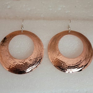 May include: A pair of large, round, copper earrings with a hammered texture. The earrings have a simple, circular design and are a warm, reddish-brown color.