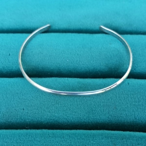 May include: A simple silver bangle bracelet, shown against a teal green background.