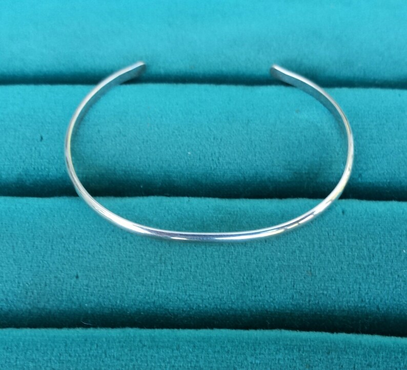 May include: A simple silver bangle bracelet, shown against a teal green background.
