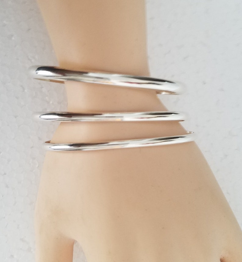 May include: Three silver bangle bracelets stacked on a model's wrist. The bracelets are smooth and shiny.