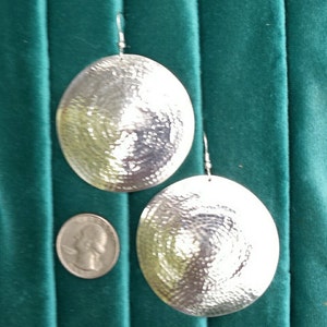 May include: A pair of large, round, silver earrings with a hammered texture. The earrings are hanging from a green background.