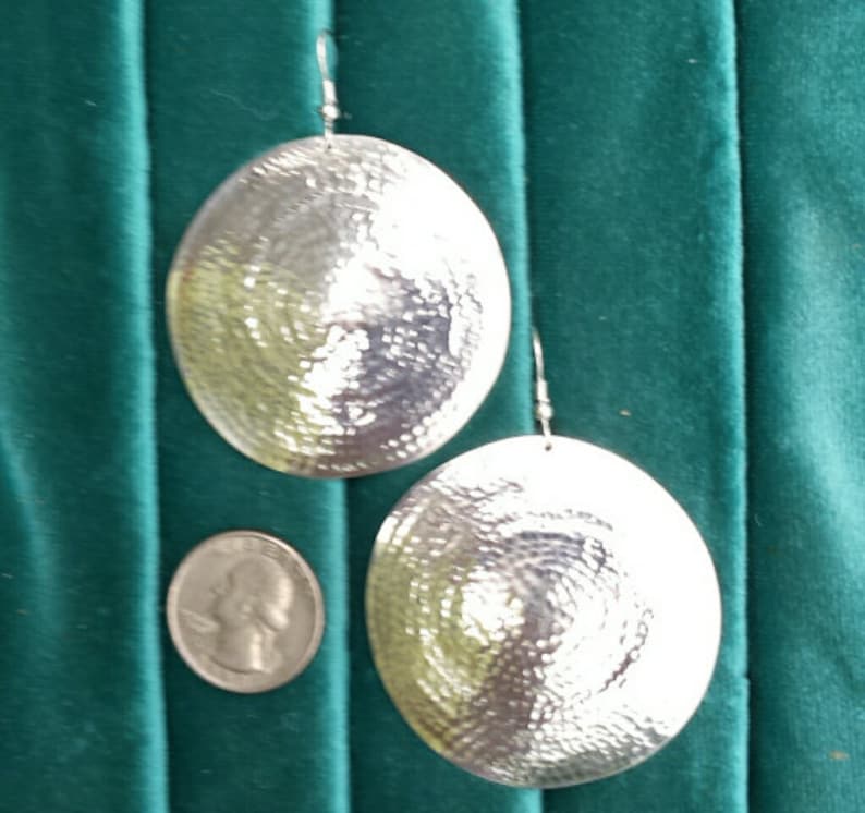 May include: A pair of large, round, silver earrings with a hammered texture. The earrings are hanging from a green background.