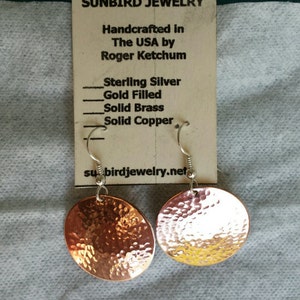 May include: A pair of hammered copper earrings with silver hooks. The earrings are round and have a textured surface. The earrings are handmade in the USA by Roger Ketchum. The card also lists the materials used: Sterling Silver, Gold Filled, Solid Brass, and Solid Copper. The website address is subirdjewelry.net.