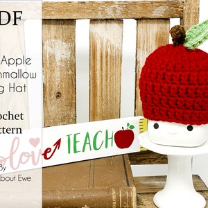 May include: A crochet pattern for a red apple-shaped mug cozy. The cozy is sitting on a white mug with a happy face. The text "VE love TEACH" is visible in the background.