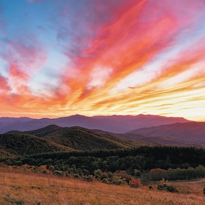 May include: A vibrant sunset over a mountain range, with a fiery orange and pink sky and a foreground of rolling hills covered in green trees.