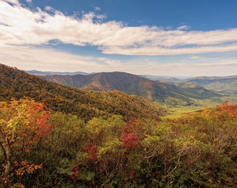 May include: A panoramic view of a mountain range with fall foliage in shades of red, orange, yellow, and green. The mountains are covered in trees and the sky is blue with white clouds.