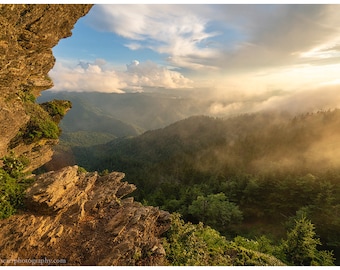 Può includere: Una vista panoramica da una scogliera rocciosa che domina una valle boscosa nebbiosa. Il sole splende attraverso le nuvole, proiettando un bagliore dorato sugli alberi sottostanti.