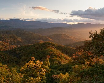 May include: A scenic landscape photograph of a mountain range at sunset. The image captures layers of rolling hills covered in lush green trees, with hints of autumn colors. The sky is a mix of blue, orange, and gray, with soft clouds.