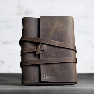 May include: A brown leather journal with a wrap-around strap closure. The journal has a distressed finish and the initials "JMT" are embossed on the lower right corner. The leather strap secures the journal, adding a rustic touch.