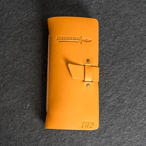 May include: A mustard-colored leather fishing log with the words "FISHING LOG" embossed on the front. The log has a buckle closure and the initials "JWP" are embossed at the bottom. The log is rectangular and appears to be made of high-quality leather.