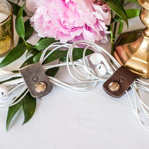May include: Two rectangular leather cord organizers in dark gray and dark brown, each featuring a small stamped heart icon and a polished gold-tone metal snap button closure, used to secure white wired earbuds in a neat, coiled arrangement.