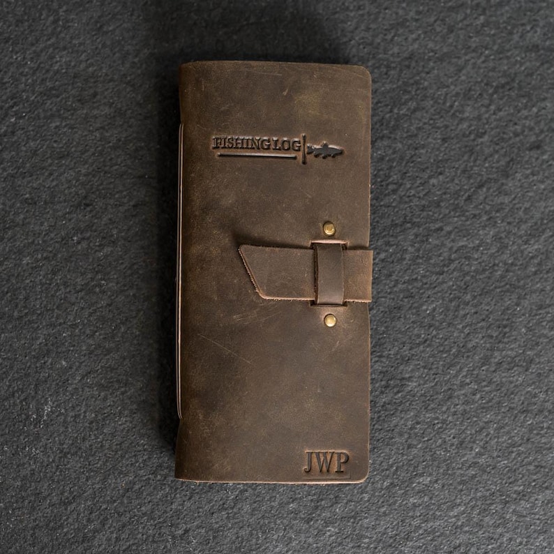 May include: A brown leather fishing log with the text "FISHING LOG" embossed on the cover. The log has a strap closure and the initials "JWP" are visible. The log is rectangular and appears to be made of distressed leather.