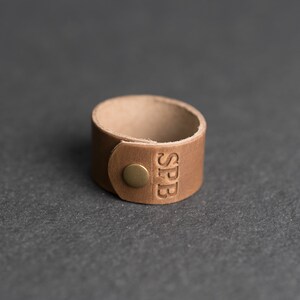 Op de afbeelding: Een beige leren manchetarmband met een ronde koperen drukknoopsluiting en de in reli&euml;f aangebrachte letters SPB aan de voorzijde. De band is gemaakt van een enkel stuk glad, lichtbruin leer met zichtbare, onafgewerkte randen en een eenvoudig ontwerp.