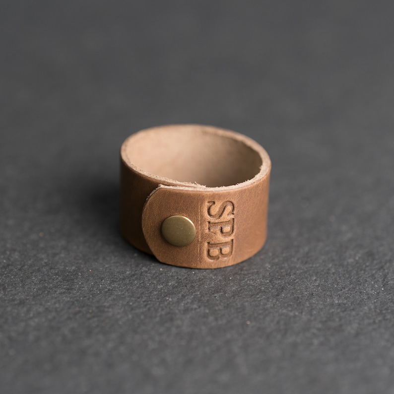Op de afbeelding: Een beige leren manchetarmband met een ronde koperen drukknoopsluiting en de in reli&euml;f aangebrachte letters SPB aan de voorzijde. De band is gemaakt van een enkel stuk glad, lichtbruin leer met zichtbare, onafgewerkte randen en een eenvoudig ontwerp.